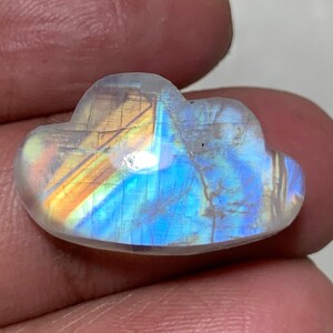 May include: A polished, cloud-shaped gemstone with a rainbow iridescence. The stone displays a spectrum of colors, including blues, oranges, and yellows. The gemstone is held in a hand, showcasing its unique shape and vibrant colors.