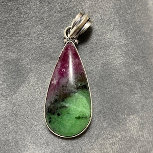 Può includere: Un ciondolo a forma di goccia con un'ansa color argento. Il ciondolo presenta una pietra lucida con una sfumatura di colori, tra cui rosso intenso, nero e verde. La pietra è incastonata in una lunetta d'argento.