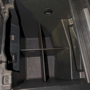 Pode incluir: Um organizador de carro de plástico preto com quatro compartimentos. O organizador está instalado no console central de um carro. O organizador tem uma porta AUX de plástico preto e uma porta USB.