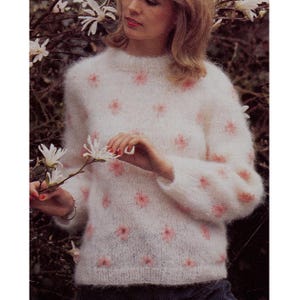 Puede incluir: Un suéter blanco y esponjoso con patrones florales rosas. El suéter tiene un cuello redondo y mangas largas. El diseño floral está espaciado uniformemente en la parte delantera y las mangas. El suéter está hecho de un material suave y texturizado.