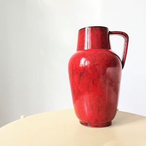 May include: A red ceramic vase with a wide mouth and a handle. The vase has a glossy finish and a speckled texture.
