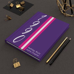 Puede incluir: Cuaderno morado con una franja blanca y rosa y el texto "0901" en blanco. El texto "Ashley Ruiz Alpha Chapter" está impreso en blanco en la parte inferior del cuaderno.