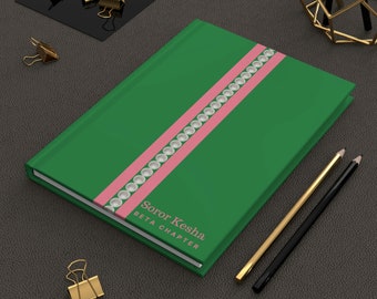 May include: A green notebook with a pink stripe and white pearls. The notebook has the text "Soror Kesha Beta Chapter" printed on the front.