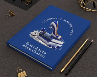 Puede incluir: Cuaderno azul con el texto "Standing on Business Since 1920" y un zapato de tacón azul sobre libros. El texto "Soror Adams Alpha Chapter" también está en el cuaderno.