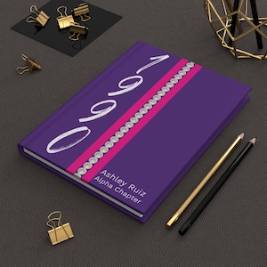 Peut inclure: Un carnet violet avec une bande rose et des accents de perles blanches. Le carnet porte le numéro "0901" écrit en blanc sur la couverture. Le texte "Ashley Ruiz Alpha Chapter" est imprimé sous le numéro.