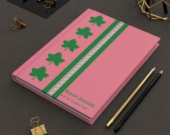 May include: A pink notebook with a green and white design. The notebook has the text "Soror Jasmin BETA CHAPTER" on the front.