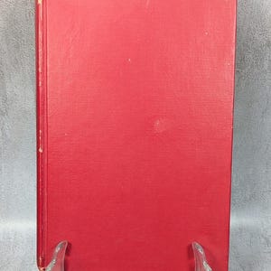 May include: A vintage hardcover book with a solid red cover. The book is displayed on a clear plastic stand. The edges of the book show signs of wear, indicating its age. The cover is plain with no visible text or design.