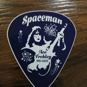 May include: A dark blue guitar pick featuring a white illustration of Ace Frehley, the Spaceman, playing a guitar. The pick has the text "Spaceman", "Ace Frehley", and the years "1951-2025".