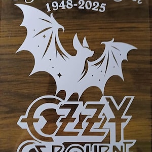 May include: White graphic design on a brown background. The design includes a bat with spread wings, the text "Flying High Again 1948-2025", and the name "Ozzy Osbourne" in a stylized font. The design is likely for a t-shirt or sticker.