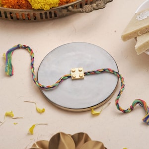 May include: A Rakhi bracelet featuring a gold-toned square charm with four raised circular studs, attached to a multi-colored braided thread made of twisted red, yellow, green, and blue fibers, with loose tassel ends on both sides of the charm.