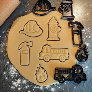 Könnte beinhalten: Ein Set aus neun schwarzen Ausstechern in Form verschiedener Feuerwehr-Motive, darunter ein Feuerwehrauto, ein Hydrant, eine Feuerwehrhaube, ein Feuerlöscher und eine Flamme. Die Ausstecher sind auf einer mit weißem Mehl bedeckten Oberfläche angeordnet.