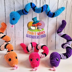 May include: A selection of crocheted snake toys in vibrant colours. The snakes are in shades of blue, orange, pink, and purple, with black and white detailing. Each snake has a small tongue and black eyes. The image includes the text "Sarah's Wooly Wonderland."