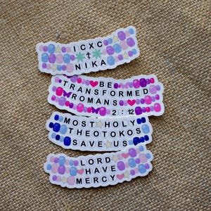 Könnte beinhalten: Fünf bunte Aufkleber mit religiösem Text und Symbolen. Die Aufkleber sind mit kleinen, bunten Perlen verziert. Der Text auf den Aufklebern lautet: "ICXC NIKA", "BE TRANSFORMED ROMANS 2:12", "MOST HOLY THEOTOKOS SAVE US", und "LORD HAVE MERCY".