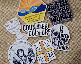 May include: A set of nine stickers with various designs and sayings. The stickers are printed on white paper with black, blue, yellow, orange, and red ink. The stickers include a sunset scene, a skull, a chalice, a cross, and a saint. The text on the stickers includes "Taste and See", "Death to the World", "Counter Culture", "St. Michael the Archangel", "IC XC NIKA", and "It is Later Than You Think".