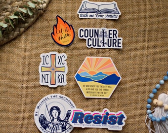 May include: A set of colorful stickers with religious and inspirational messages. The stickers feature a cross, a sunrise, a flame, a book, and a figure of St. Michael the Archangel. The text on the stickers includes "Let it Shine", "Teach me Your Statutes", "Counter Culture", "IC XC NI KA", "He who gives you the day will also give you the things necessary for the day", "Resist", "St. Michael the Archangel Pray to God For".