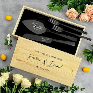 May include: A wooden box with a black engraved inscription that reads "OUR WEDDING MEMORIES Kristine & Daniel MAY 18, 2024". The box contains a black cake knife and two black forks.