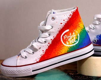 May include: White high-top canvas sneakers with a rainbow paint splatter design. The sneakers have a white logo that says "Pintar i Fet" on the side.