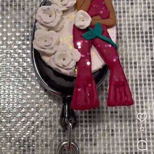 May include: A red and white badge reel featuring a woman with long black hair singing into a microphone. The woman is surrounded by white roses with glitter accents.