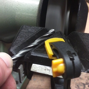 Peut inclure: Un gabarit de meulage de foret noir avec une pince jaune, maintenant un foret en place contre une meule.