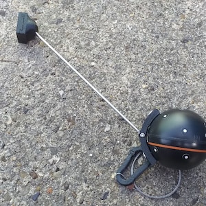 May include: A black and orange compass with a silver rod attached to a black clip. The compass is lying on a grey concrete surface.