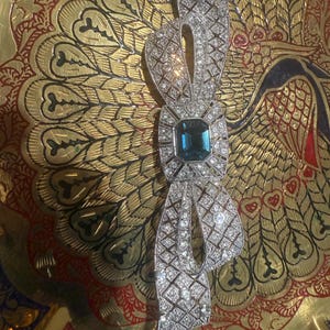 May include: A silver-toned brooch in a bow shape, encrusted with small, sparkling crystals. The center features a square, teal-colored gemstone. The brooch is set against a gold and red decorative surface.