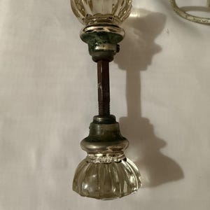 May include: A vintage glass door knob with a fluted design. The knob has a clear, faceted glass top and bottom, with a tarnished metal base and a long, threaded rod. The knob is likely from an antique door.