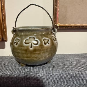 May include: A small, round, brown ceramic cauldron with a glossy finish, featuring embossed shamrock designs around the exterior. The pot has three short feet, two side handles, and a thin, dark metal wire handle arched over the top opening.