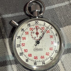 May include: A silver-toned Compass stopwatch with a white face and red numerals. The watch features a large, easy-to-read dial with a second hand and a smaller inner dial. The stopwatch is marked "SWISS MADE".