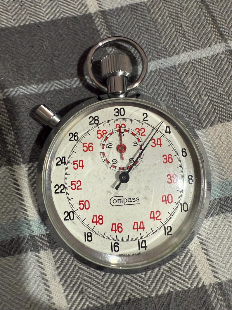 May include: A silver-toned Compass stopwatch with a white face and red numerals. The watch features a large, easy-to-read dial with a second hand and a smaller inner dial. The stopwatch is marked "SWISS MADE".