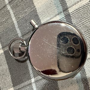May include: A shiny, silver stopwatch with a ring and a button. The stopwatch is round and has a reflective surface, showing a distorted reflection. The background is a gray and white checkered pattern.
