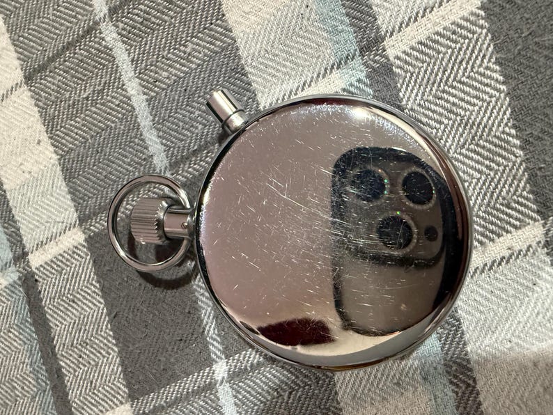 May include: A shiny, silver stopwatch with a ring and a button. The stopwatch is round and has a reflective surface, showing a distorted reflection. The background is a gray and white checkered pattern.