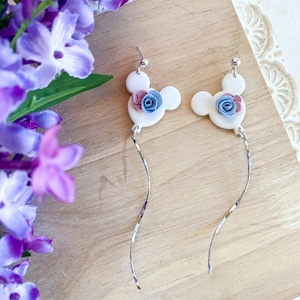 May include: A pair of white dangle earrings with a blue and pink flower design. The earrings are shaped like Mickey Mouse ears.