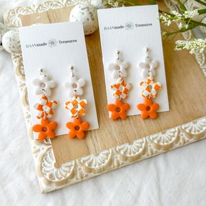May include: Two pairs of orange and white flower dangle earrings on a white card with the text "HAANmade Treasures".
