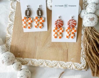 May include: Two pairs of handmade earrings displayed on white cards. One pair features brown football-shaped tops with orange and white checkered bows. The other pair has orange basketball-themed tops with orange and white checkered bows. The cards read "HAANmade Treasures."