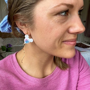 May include: A pair of white dangle earrings with a blue and pink flower design. The earrings are shaped like a cartoon mouse head.