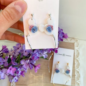 May include: White ceramic earrings with a floral design. The earrings feature a cluster of three flowers in shades of pink, blue, and purple. The earrings are attached to silver-toned, thin, and slightly twisted wires.