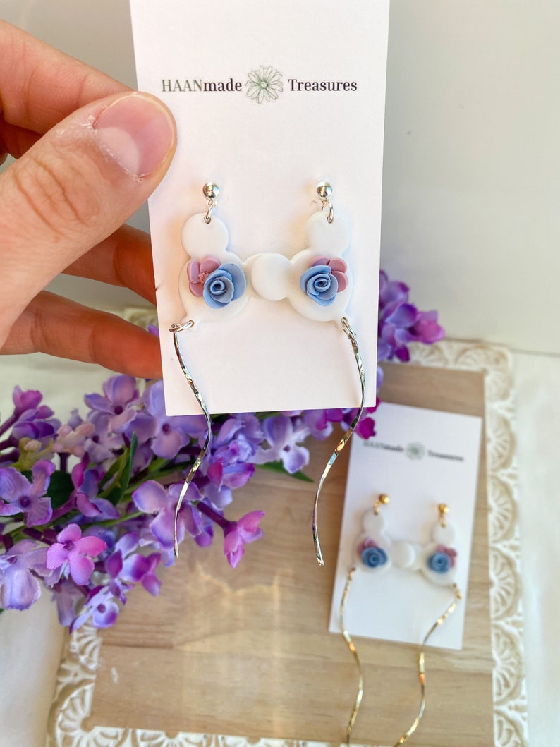 May include: White ceramic earrings with a floral design. The earrings feature a cluster of three flowers in shades of pink, blue, and purple. The earrings are attached to silver-toned, thin, and slightly twisted wires.