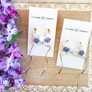 May include: A pair of white dangle earrings with blue and pink flower accents. The earrings are on a white card with the text "HAANmade Treasures".
