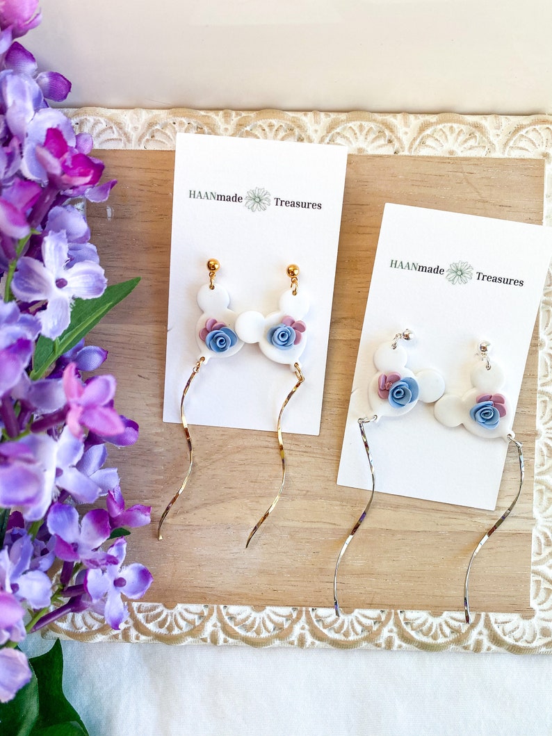 May include: A pair of white dangle earrings with blue and pink flower accents. The earrings are on a white card with the text "HAANmade Treasures".