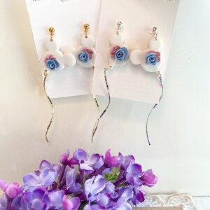 May include: A pair of white ceramic dangle earrings with blue and pink flower accents. The earrings are on a white card with the text "HAANmade Treasures".