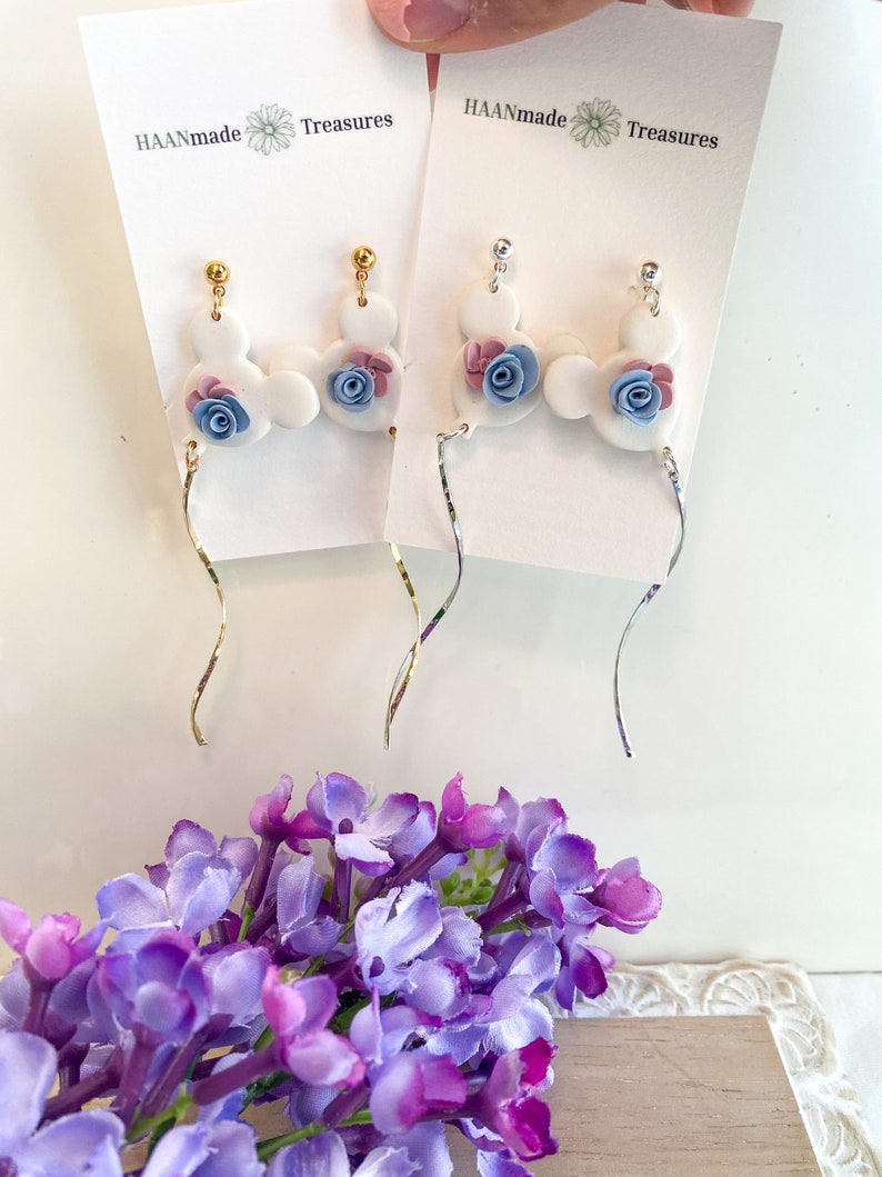 May include: A pair of white ceramic dangle earrings with blue and pink flower accents. The earrings are on a white card with the text "HAANmade Treasures".