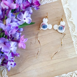 May include: A pair of white dangle earrings with a gold chain. Each earring features a white Mickey Mouse head with a blue and pink flower.