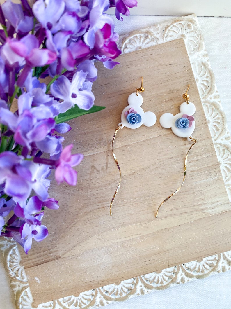 May include: A pair of white dangle earrings with a gold chain. Each earring features a white Mickey Mouse head with a blue and pink flower.