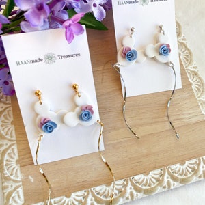 May include: Two pairs of dangle earrings with white clay charms shaped like Mickey Mouse ears with blue and pink flowers. The earrings are displayed on white cards with the text "HAANmade Treasures".