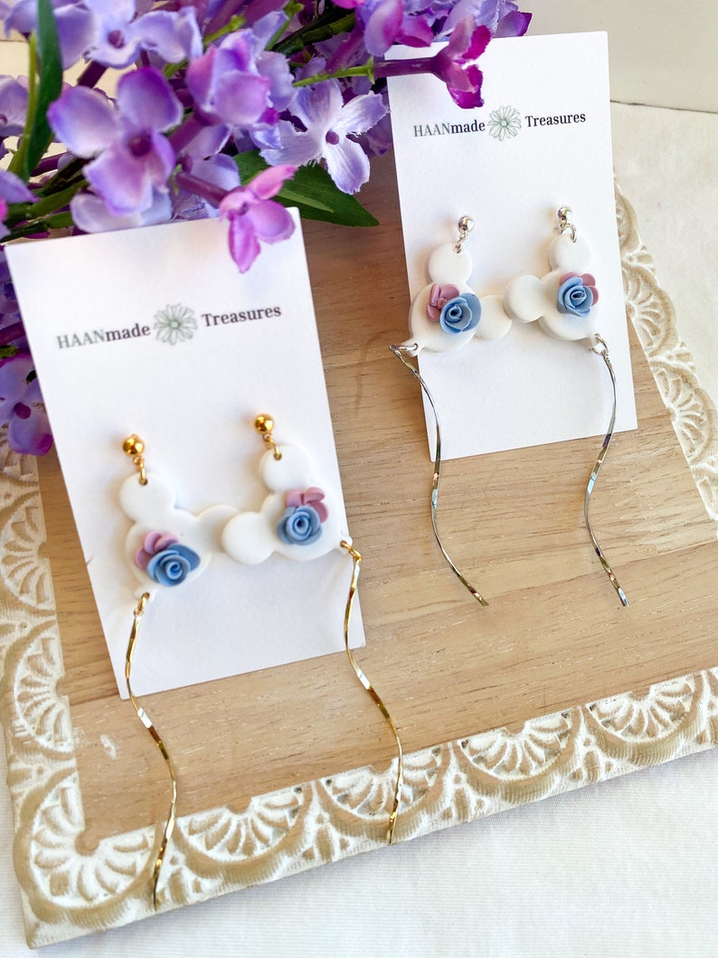 May include: Two pairs of dangle earrings with white clay charms shaped like Mickey Mouse ears with blue and pink flowers. The earrings are displayed on white cards with the text "HAANmade Treasures".