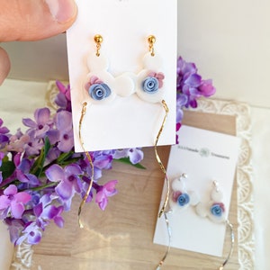 May include: White ceramic earrings with a blue and pink flower design. The earrings are dangle style with a gold chain.