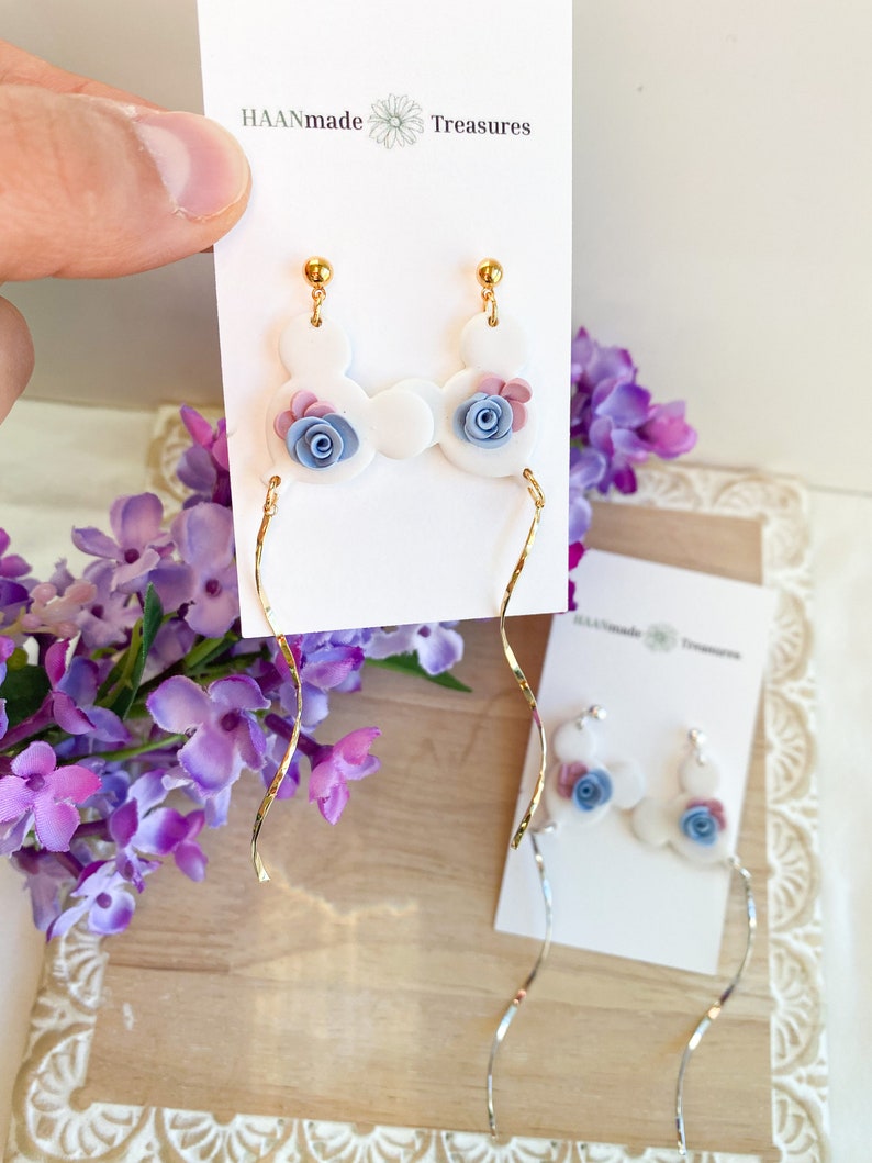 May include: White ceramic earrings with a blue and pink flower design. The earrings are dangle style with a gold chain.