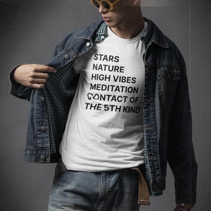 May include: A white t-shirt with black text that reads "Stars, Nature, High Vibes, Meditation, Contact of... The 5th Kind". The person is wearing a denim jacket and jeans.