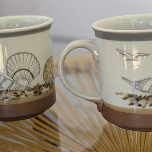 May include: Two ceramic mugs with a beach theme. The mugs are white with a brown base and a gray rim. Each mug features illustrations of seashells, a starfish, and a flying seagull.