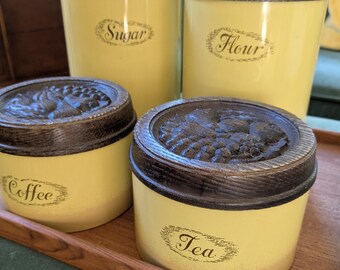 May include: A set of four yellow kitchen canisters with dark brown lids. The canisters are labeled "Sugar", "Flour", "Coffee", and "Tea" in an elegant script. The lids have a detailed embossed design. The canisters are displayed on a wooden tray.